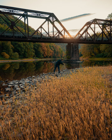 A back view of a man throwing stone in river under a bridge in autumnの写真素材