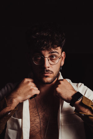 A portrait of a young sexy Hispanic man with glasses and beard wearing a white shirt, black...