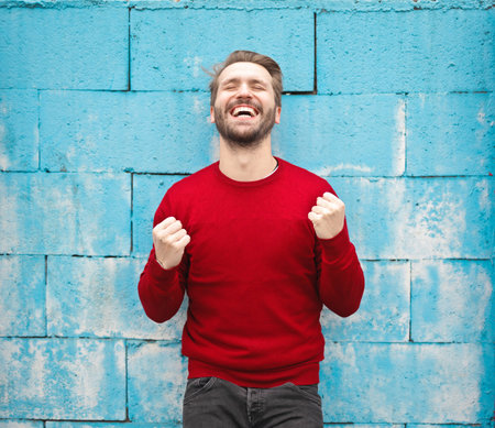 A Caucasian young man celebrating victory with his fists up on background of a blue wallの写真素材
