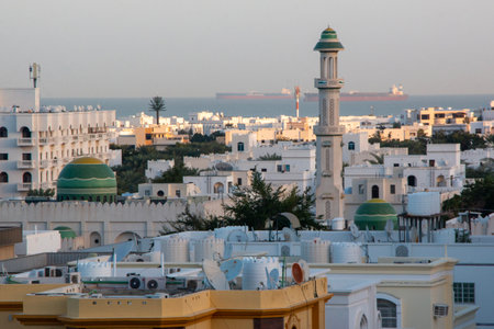 A beautiful shot of a white Mosque with green domes in the neighborhood with a sailing ships in the sea in the background on a sunny dayの写真素材