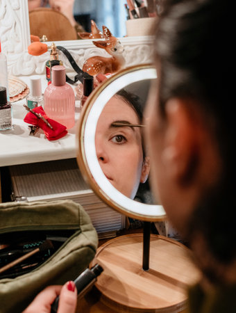 Selective focus Portrait of young woman in mirror doing her make-up. Woman using eyebrow brush in...