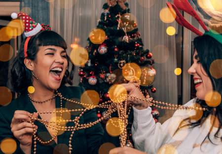 Two beautiful happy young women decorating christmas tree on christmas eve. Bokeh lights overlay...