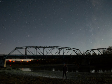 A scenic view of a person standing on bank of a river against a bridge, looking at starry night skyの写真素材