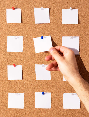 A vertical shot of a person's hand holding a blank paper note with tack on a notes boardの写真素材