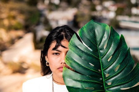 An elegant woman dressed in a white suit covering half her face with a green leaf on a mountain at sunsetの写真素材