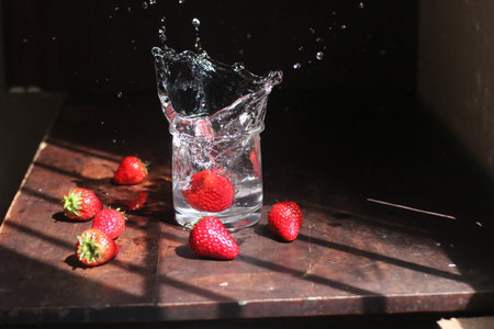 A closeup of a strawberry falling with splash on a cup of water next to strawberries on a wooden...