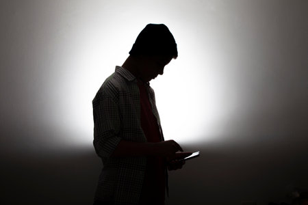 A silhouette view of a young man using his smartphone on a lighting gray backgroundの写真素材