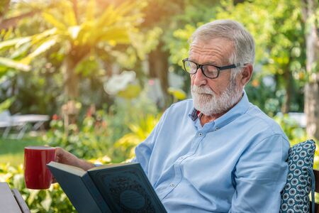 Senior elderly man reading book drinking mug of coffee in gardenの写真素材