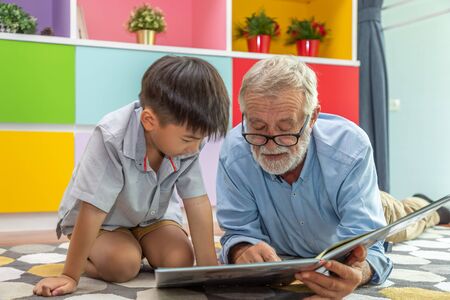 Happy boy grandson reading book with old senior man grandfather at homeの写真素材