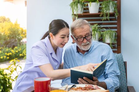 Senior elderly man reading book with nurse during breakfast in garden at nursing homeの写真素材