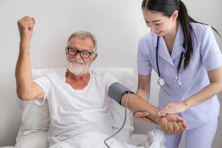 Happy nurse checking and measuring elderly man blood pressure in bedroom at nursing home, rise fist upの写真素材