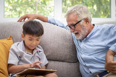 Happy boy grandson playing game on tablet with old senior man grandfather at homeの写真素材