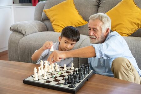 Happy boy grandson playing chess with old senior man grandfather at homeの写真素材
