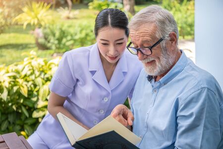 Senior elderly man reading book with nurse during breakfast in garden at nursing homeの写真素材
