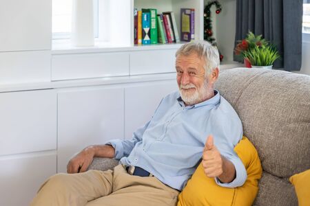 Relax happy senior old man eldery sitting on comfortable sofa in living room thumb up looking cameraの写真素材