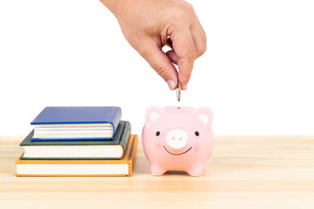 Person hand putting depositing money into pink piggy bank on wooden desk with books stack on white background, concept saving for education and scholarshipの写真素材