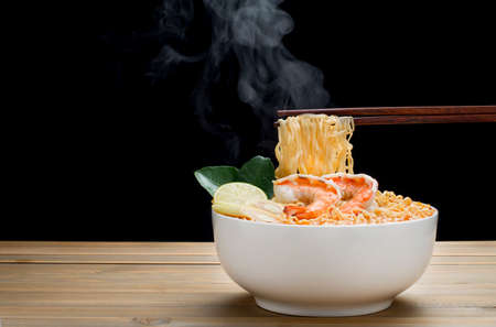 Use chopsticks to eat delicious hot and sour Tom Yum Kung noodles. And with hot steam in a white bowl placed on a wooden table on a black backgroundの写真素材