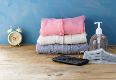Knit sweaters are stacked on a wooden table with a mask and anti-COVID-19 hand sanitizer.