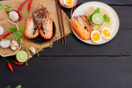 Tom Yum Goong Instant Noodle Ramen - Asian Style Thai and Chinese are spicy. placed on a black wooden table With seafood shrimp, lime, red chili, garlic, galangal, lemongrass, kaffir lime stand, top view.の写真素材