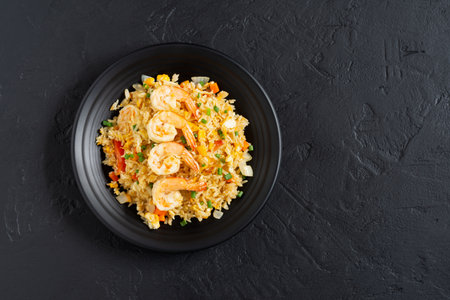 Fried rice with vegetables and sea prawns, carrots, eggs, and tomatoes. Thai Chinese food is arranged on a black plate on a black background kitchen table, top view.の写真素材