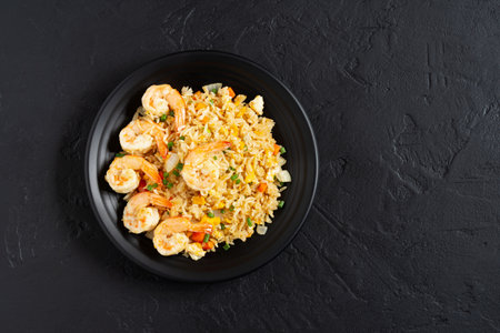 Fried rice with vegetables and sea prawns, carrots, eggs, and tomatoes. Thai Chinese food is arranged on a black plate on a black background kitchen table, top view.の写真素材