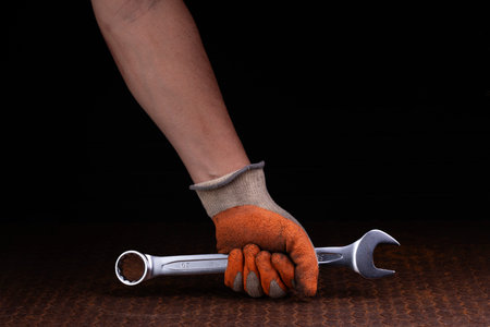 Mechanic in orange gloves holding wrench on metal table, isolated on black and dark background.