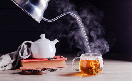 Pour hot tea into cups, herbal tea glasses are arranged on a rustic wooden table.