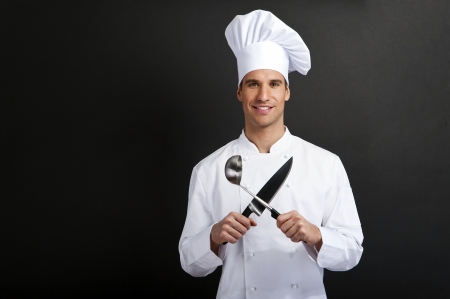 Chef cook against dark background smiling with hat holdinf spoon and knifeの写真素材