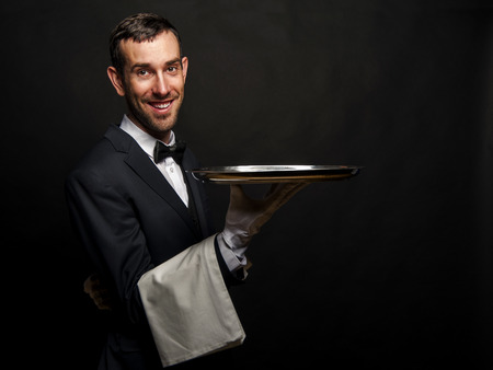 Waiter in black suit holding tray over black background.