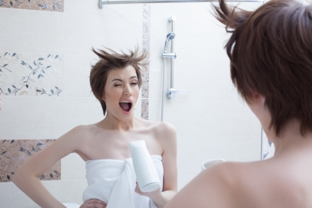 Cheerful girl posing with hairdryer near mirror in bathroomの写真素材