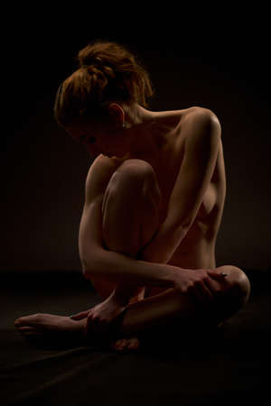 Slim naked woman sitting in darkness in studioの写真素材