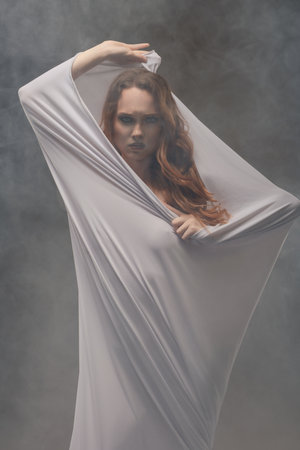 Female model draped in sheer fabric poses dramatically in a misty studio settingの写真素材