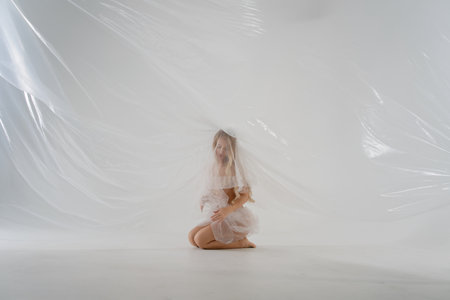 Blonde woman in soft attire kneels in a bright studio with drapesの写真素材