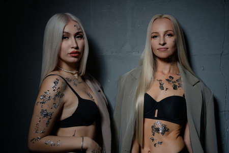 Two models in silver body art pose in stylish outfits on a dark backdropの写真素材