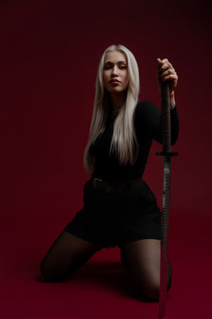 Woman in black outfit poses with sword against a red background in a dramatic settingの写真素材