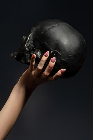 Womans hand with decorated fingernails holding a black skull against a dark gray background in a studioの写真素材