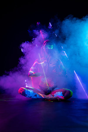 Woman in UV body paint and cyber glasses sits in a meditative pose amidst purple smoke and laser...