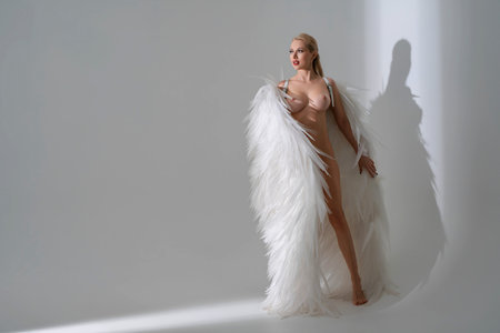 Ethereal blonde woman wearing large white angel wings stands in a minimalist studio.の写真素材