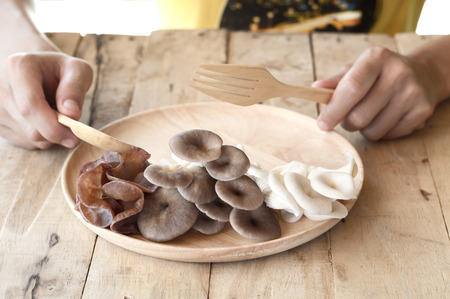 side view of woman eating different edible mushroom in wooden dish with spoon and fork on wood tableの写真素材