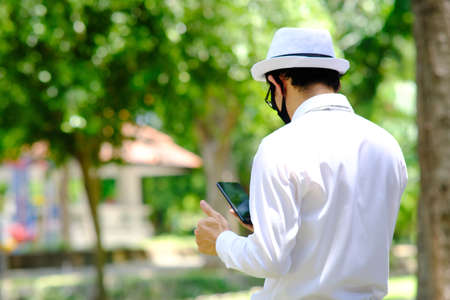 Asian man wearing a white shirt and hat using a tablet in the park