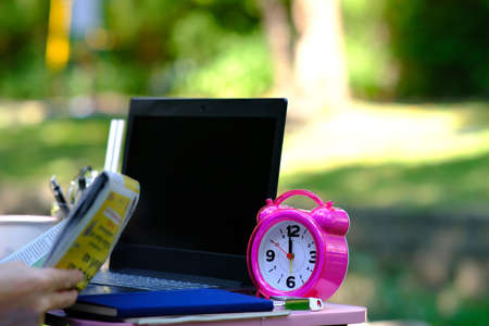 Close-up of a woman's hand holding a laptop and a pink alarm clock on a wooden table in the park.