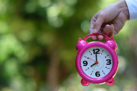 Hand holding pink alarm clock on blurred nature background. Time management concept.