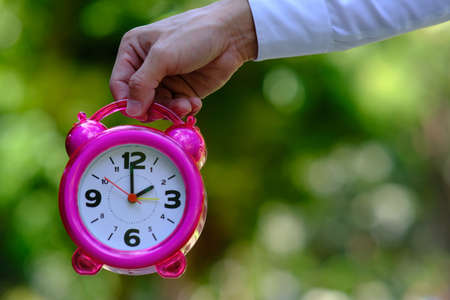 Hand holding pink alarm clock on blurred green background. Time management conceptの写真素材