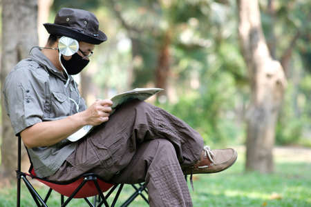 Asian man wearing face mask reading a book while sitting in the park.の写真素材