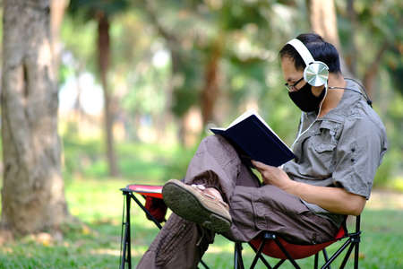 Young Asian man reading a book while wearing face mask in the park.