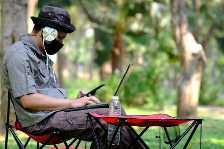 Asian man wearing a mask and using a laptop in the park.