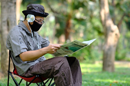 Asian man wearing face mask reading newspaper while sitting in the park.