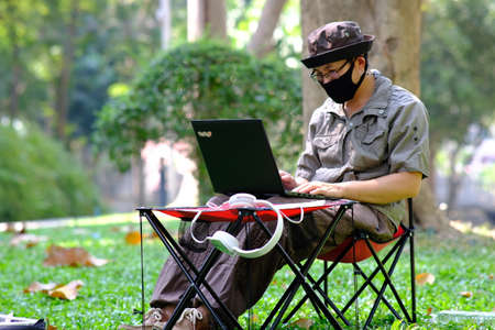 A man in a protective mask works on a laptop in the park.