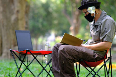 A man in a protective mask works on a laptop in the park.