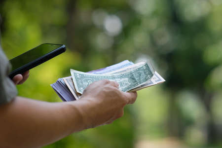 A man's hand holding a smartphone and counting money in the parkの写真素材